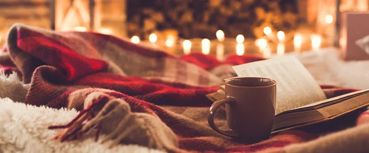 Gemütliche Szene mit einem aufgeschlagenen Buch, einer Tasse und einer rot-karierten Decke. Warme Lichter funkeln im Hintergrund und schaffen eine behagliche Atmosphäre.