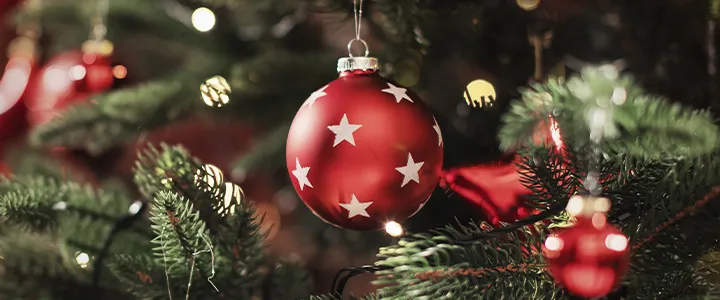 Rotes Weihnachtsornament mit weißen Sternen, das an einem geschmückten Baum hängt, umgeben von Lichtern und grünen Zweigen.