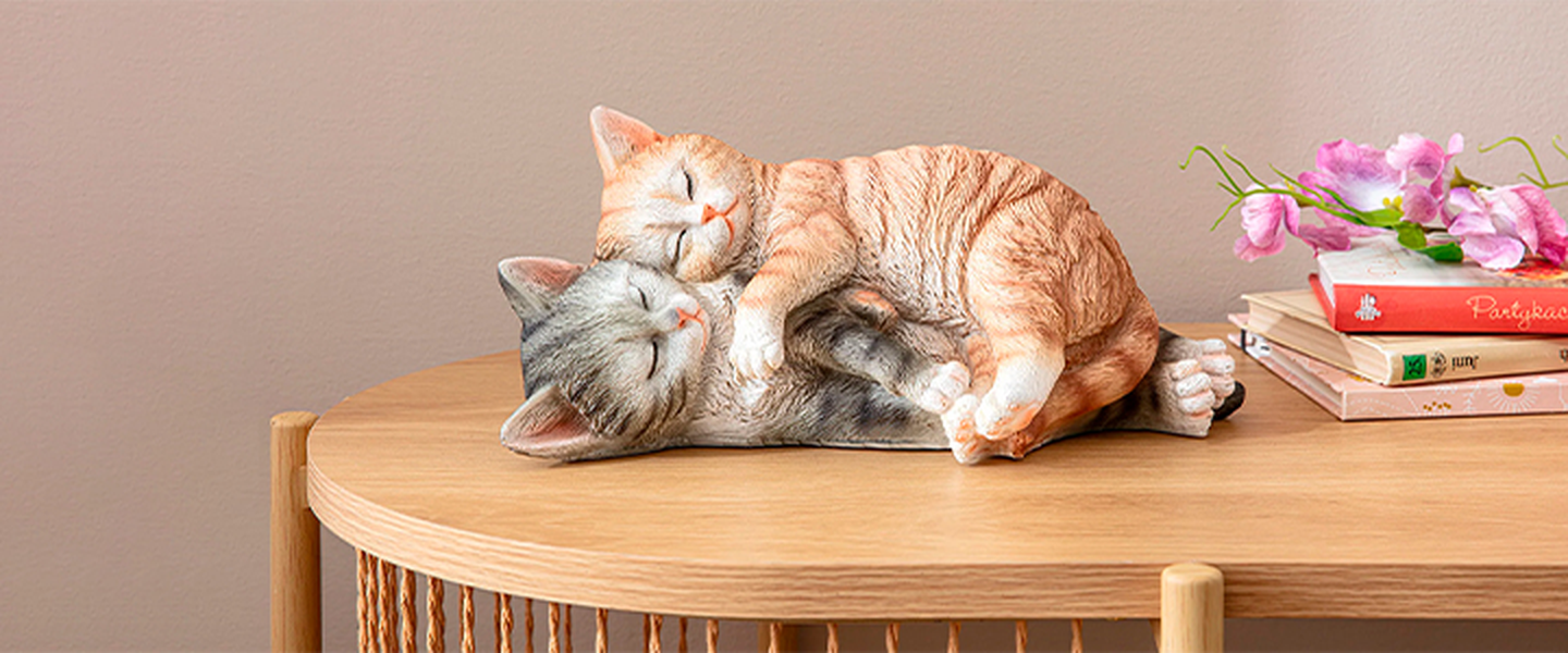 Zwei realistische Katzenfiguren, eine orangefarbene und eine graue, schlafen zusammengerollt auf einem Holztisch neben gestapelten Büchern und rosa Blumen.