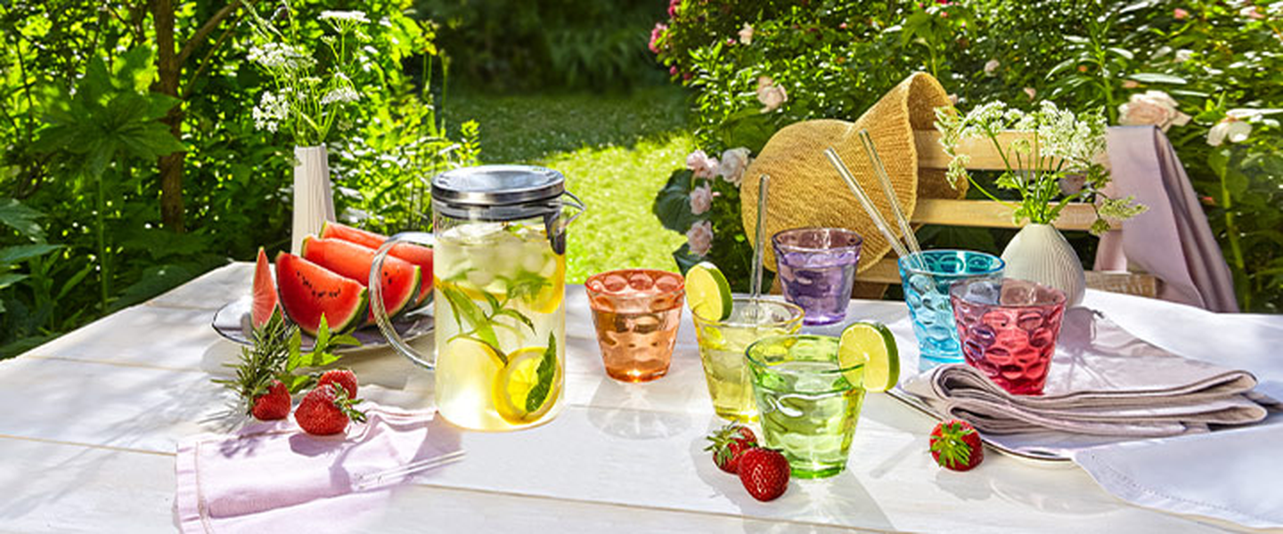 Ein sonniger Gartentisch, gedeckt mit bunten Gläsern, einem Krug Zitronenwasser, frischen Wassermelonenscheiben, Erdbeeren und Sonnenhüten auf Stühlen, umgeben von Grün.