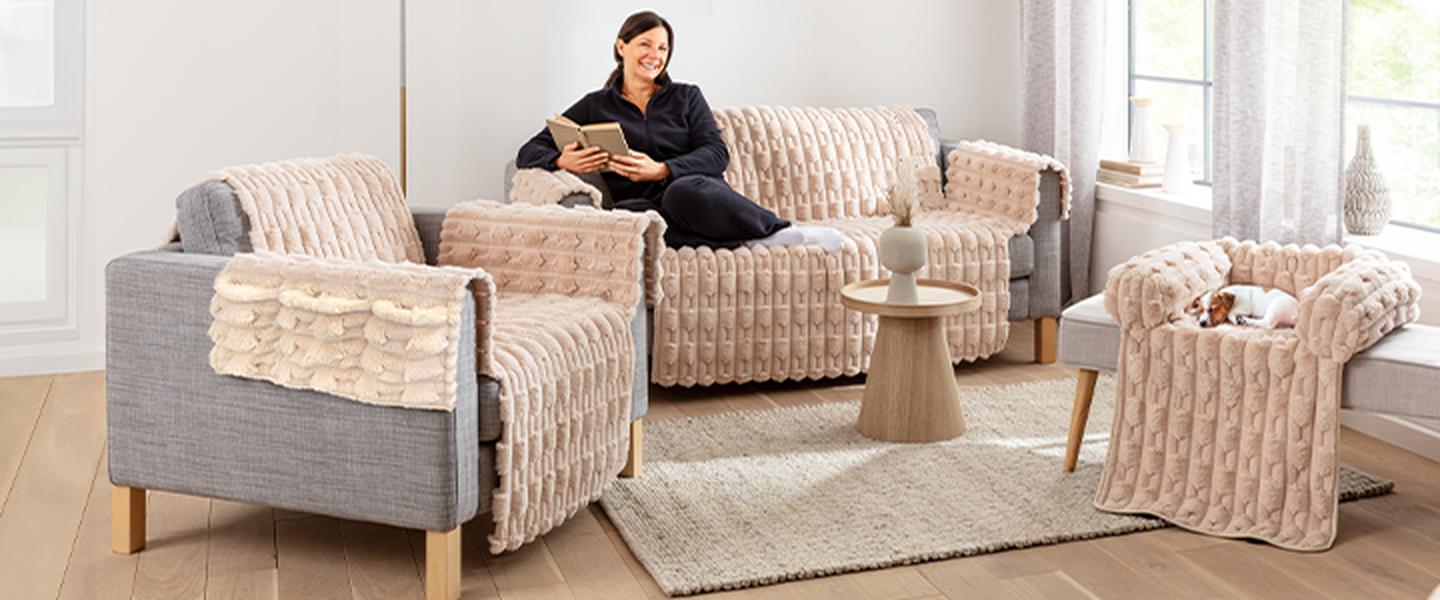 Eine Frau sitzt auf einem gemütlichen, beigen Sofa und liest ein Buch. Ein kleiner Hund schläft auf einem passenden Sessel. Heller Raum mit großen Fenstern.