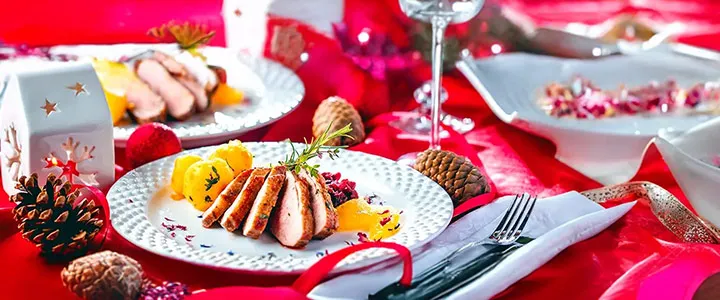 Festlich gedeckter Tisch: weißer Teller mit aufgeschnittenem Bratenfleisch, Orangenspalten und Kräutern, Tannenzapfen und rote Tischdecke mit Weinglas.