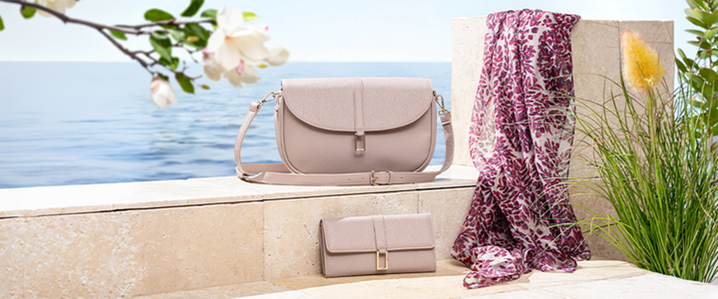 Eine beige Handtasche und eine Geldbörse sowie ein geblümter Schal liegen auf einem Felsvorsprung am Meer, umgeben von Grün und blühenden Blumen.