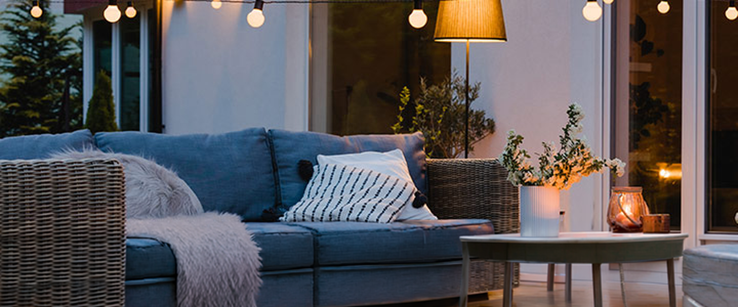 Gemütliche Terrasse mit Korbmöbeln, Kissen und Lichterketten. Ein kleiner Tisch mit Kerzen sorgt für eine warme Atmosphäre.