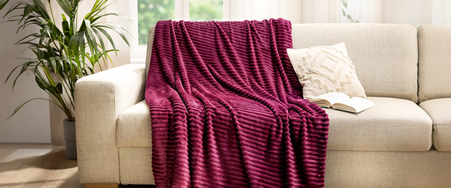 Ein beiges Sofa mit einer violetten Decke mit Strukturmuster und einem cremefarbenen Kissen, aufgestellt in einem hellen Raum mit Holzböden und einer Topfpflanze in der Nähe.