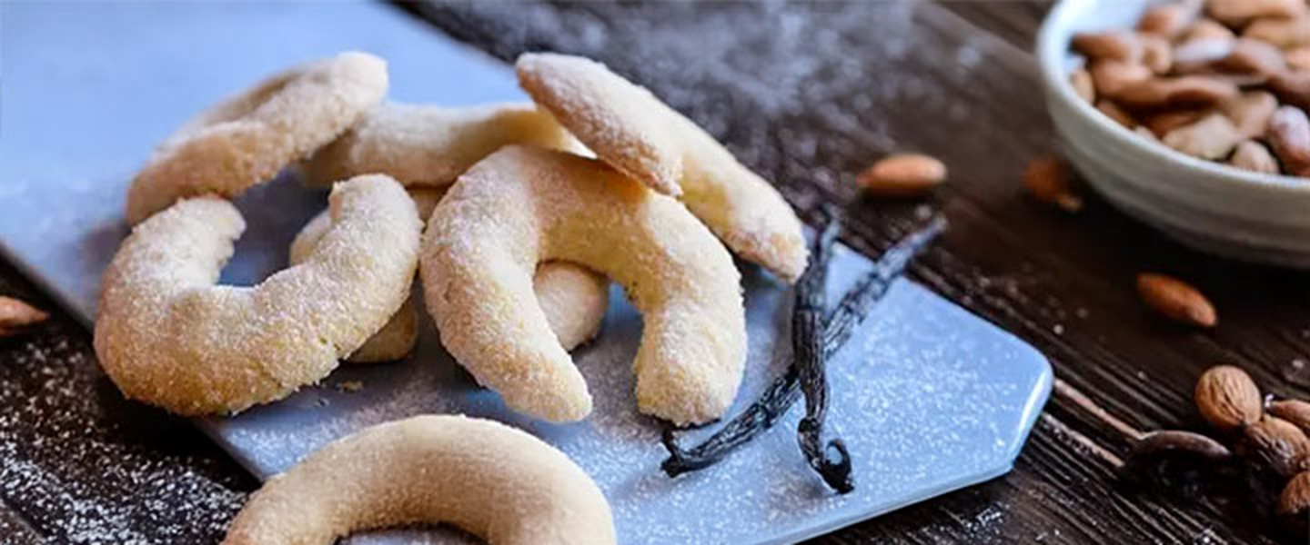 Mit Puderzucker bestäubte Mandelhörnchen auf einem blauen Brett, dazu Vanilleschoten und eine Schüssel mit ganzen Mandeln auf einem dunklen Holztisch.