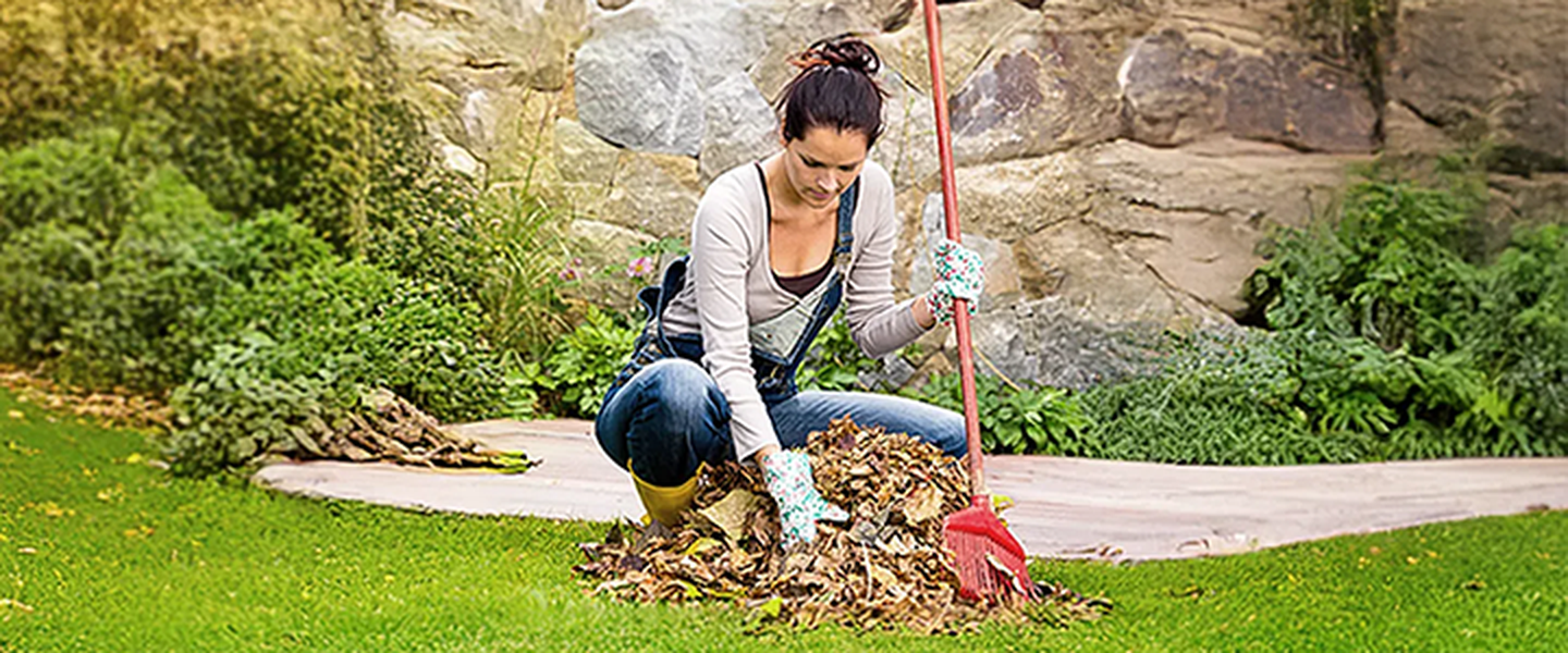 Eine Frau die im Garten vor einer Steinmauer das Laub weg macht