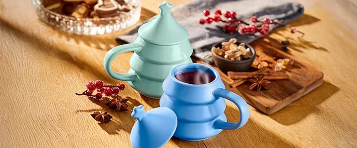 Zwei bunte Tassen mit Deckeln auf einem Holztisch, umgeben von Gewürzen, Nüssen und einer Glasschale mit Keksen. Eine gemütliche, festliche Umgebung.