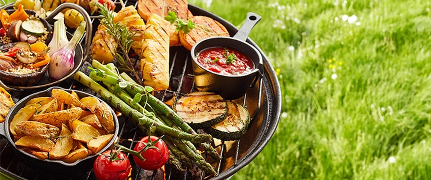 Ein Grill mit verschiedenen Gemüsesorten, Kartoffeln und Soße vor einer Kulisse aus saftig grünem Gras.