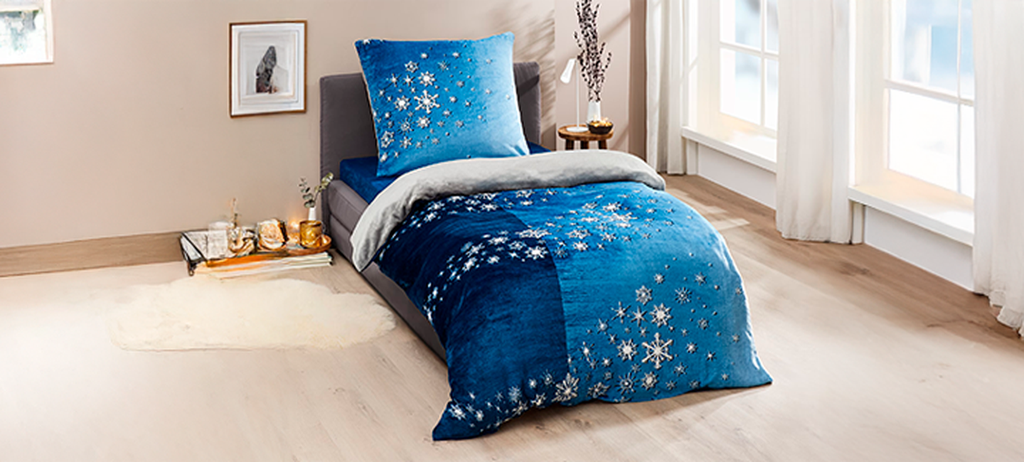 Gemütliches Schlafzimmer mit einem Bett mit blauer Bettwäsche mit Schneeflockenmuster, weichen Kissen und hellem Tageslicht durch große Fenster.
