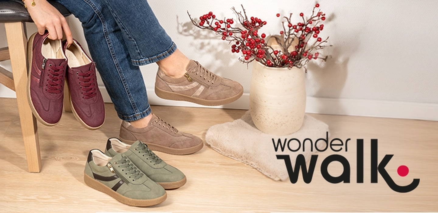Eine Person sitzt auf einem Hocker und hält burgunderrote Turnschuhe in der Hand; auf dem Boden liegen beige und grüne Turnschuhe neben einer Vase mit roten Stielen.