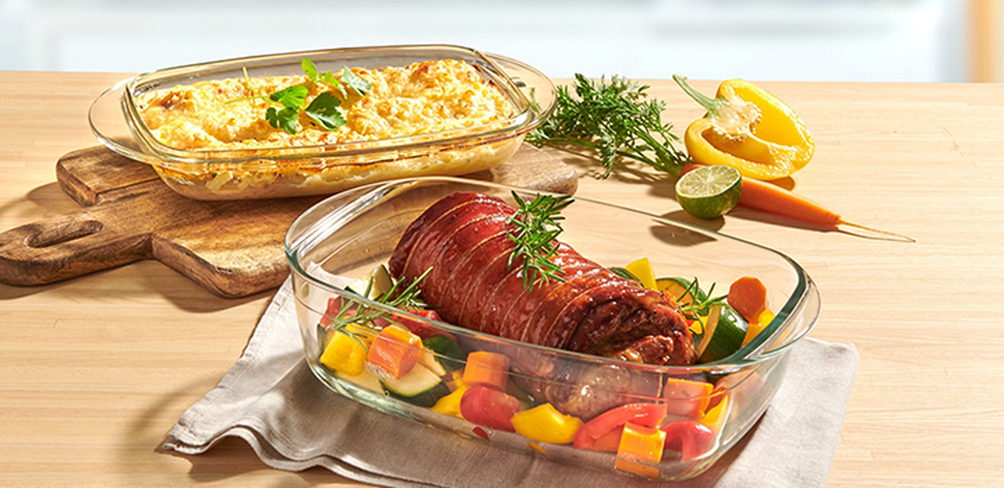 Roastbeef und Gemüse in einer Glasform neben einem überbackenen Auflauf, garniert mit Kräutern, auf einem Holztisch mit in Streifen geschnittener Paprika und Limette.
