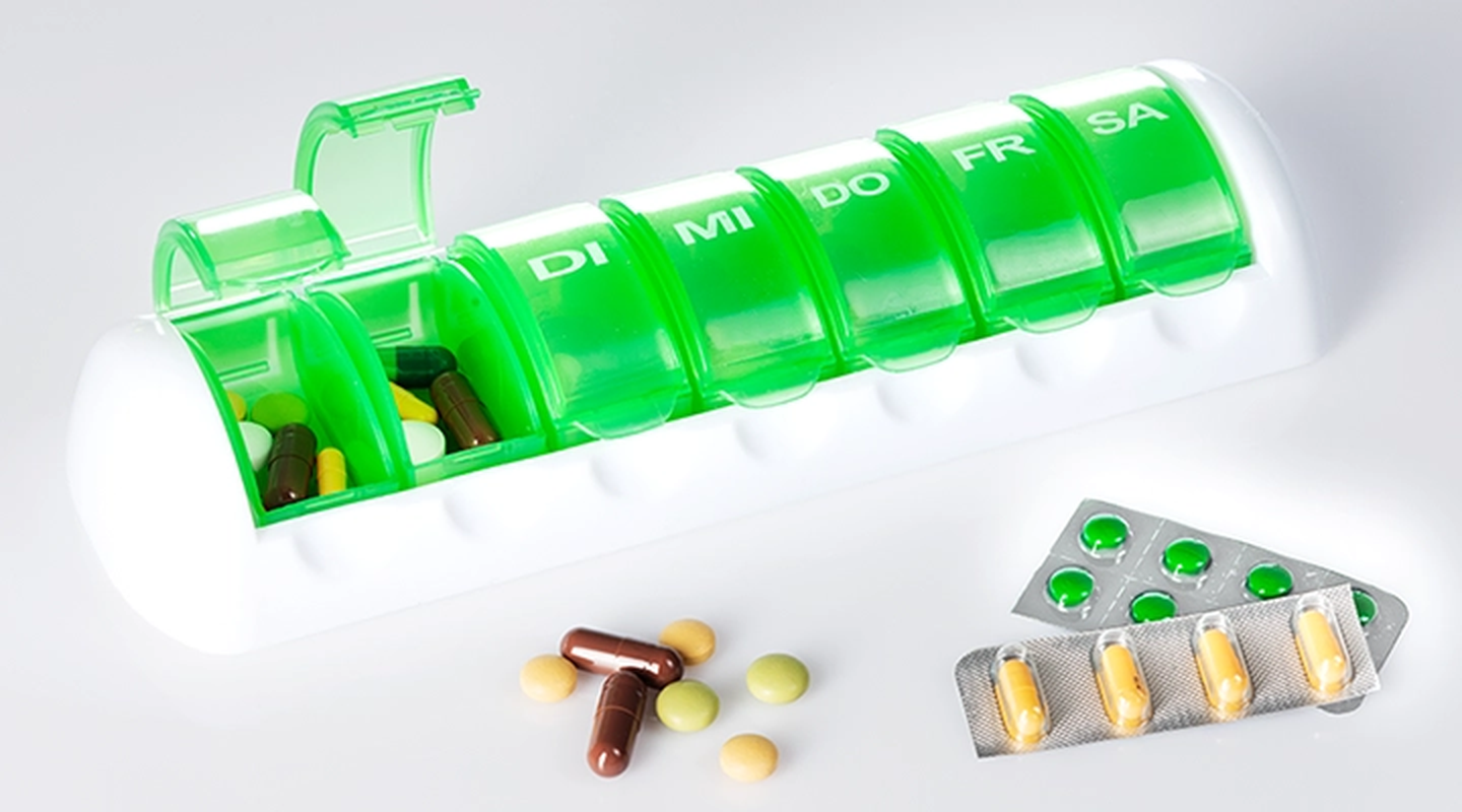 Grün-weißer Tablettenbehälter mit Fächern für die Wochentage, daneben verschiedene Tabletten und Blisterpackungen auf einer weißen Fläche.