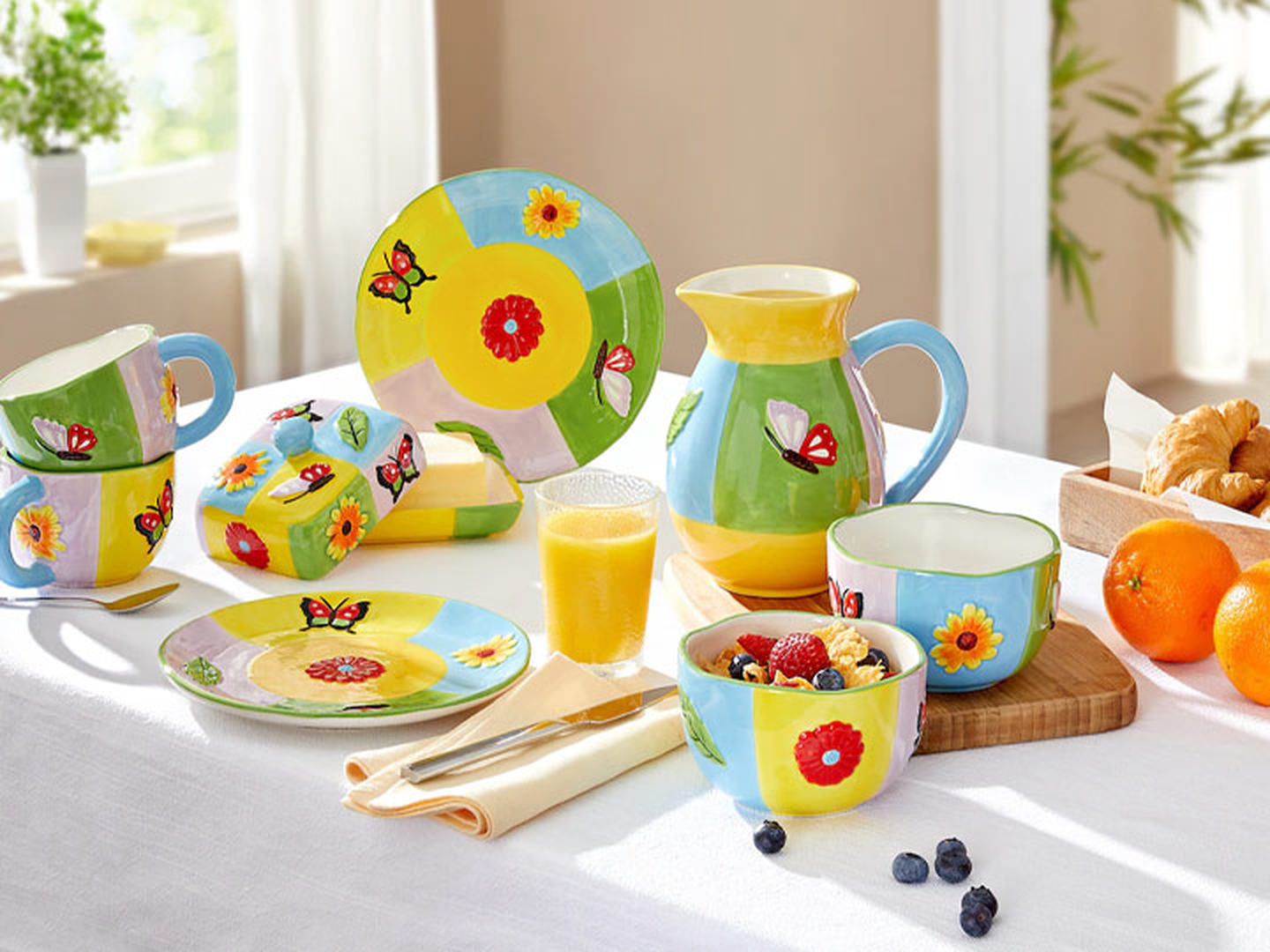 Ein farbenfrohes Frühstücksset aus Keramik mit Blumen- und Schmetterlingsmotiven, bestehend aus Tellern, Schüsseln, Bechern und einem Krug, steht auf einem weißen Tisch mit Obst und Saft.