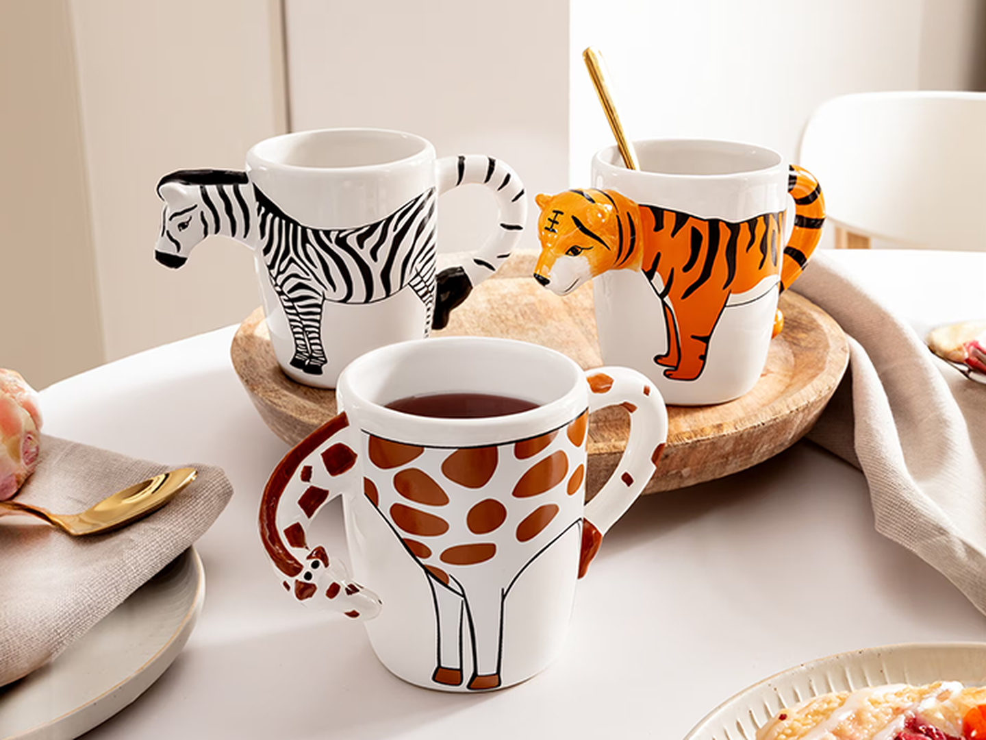 Drei Tassen mit Tiermotiven – Zebra, Tiger und Giraffe – und außergewöhnlich geformten Henkeln stehen auf einem Tisch, zusammen mit Tee und einem goldenen Löffel.