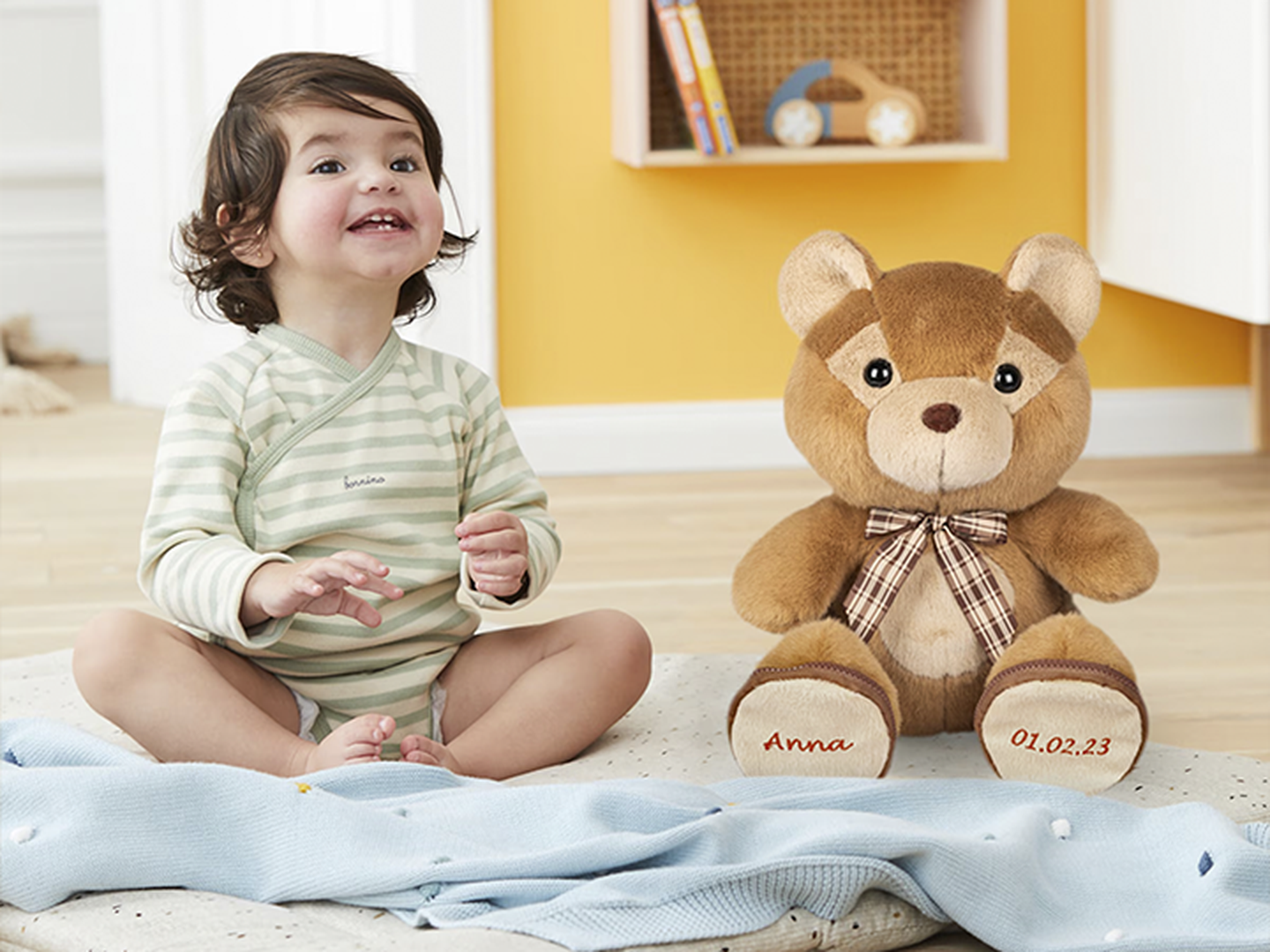 Ein lächelndes Kleinkind in gestreiftem Outfit sitzt auf einer Decke neben einem großen Teddybären mit Schleife in einem gemütlichen Zimmer mit Büchern im Regal.