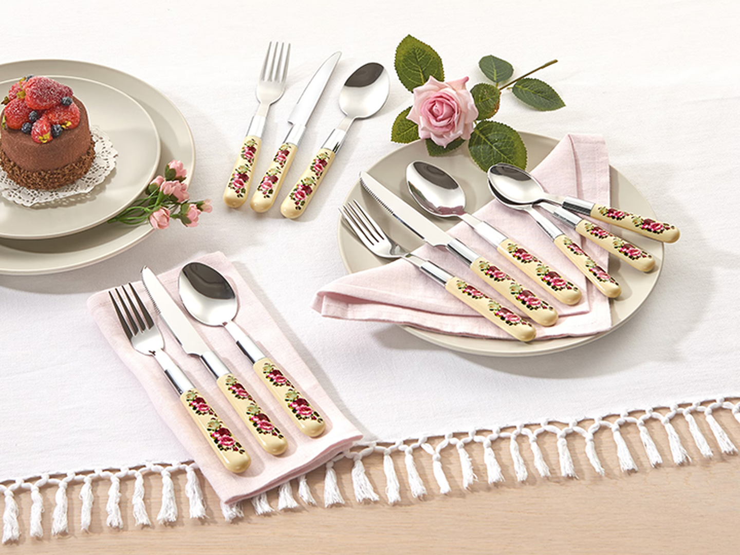Besteckset mit floralen Griffen, arrangiert auf einer weißen Tischdecke mit einer rosa Serviette, daneben ein Dessert und eine rosa Rose.