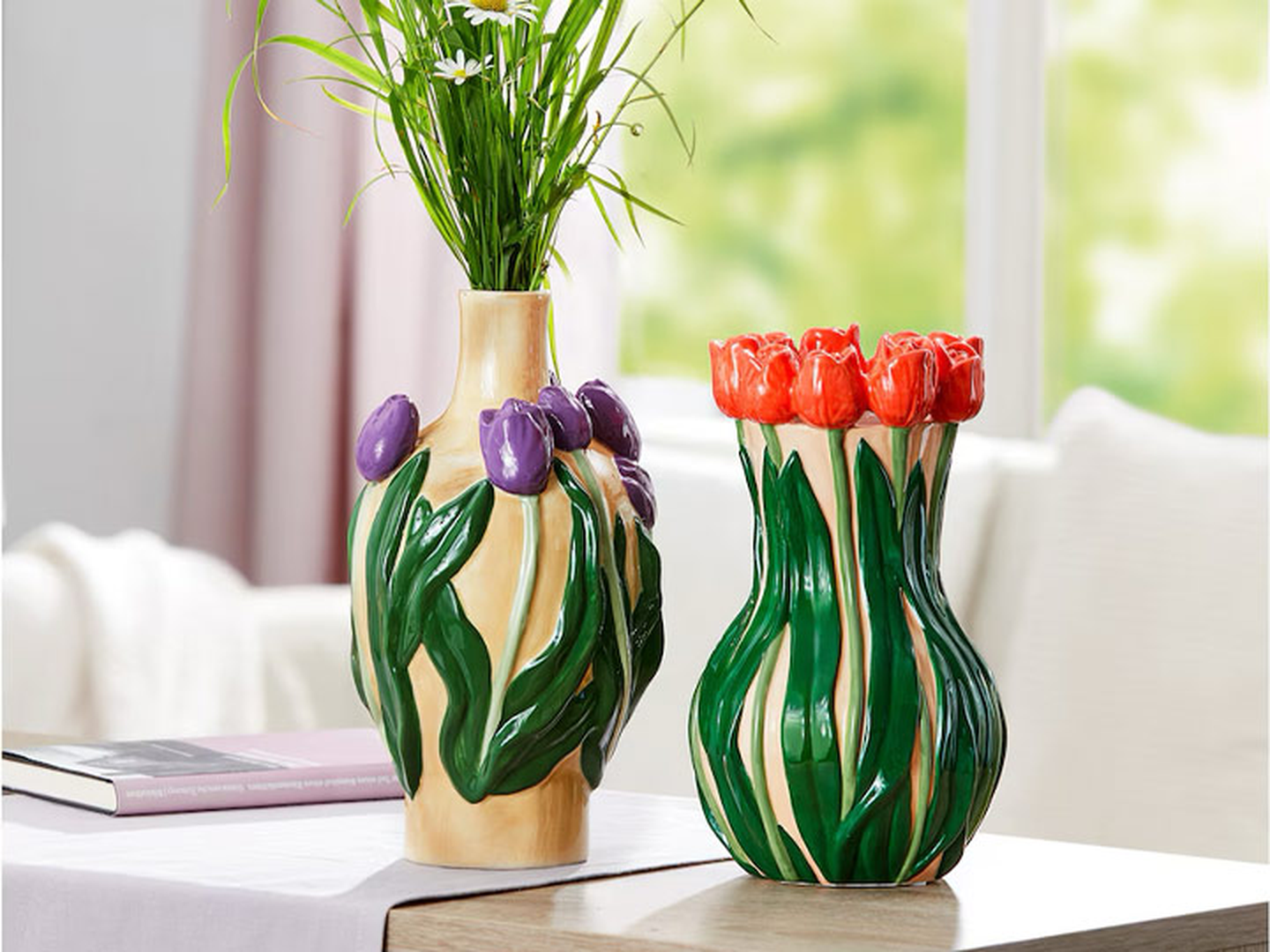 Zwei dekorative Vasen mit Tulpenmotiven, eine davon mit Blumen gefüllt, stehen auf einem Tisch in der Nähe eines Fensters.