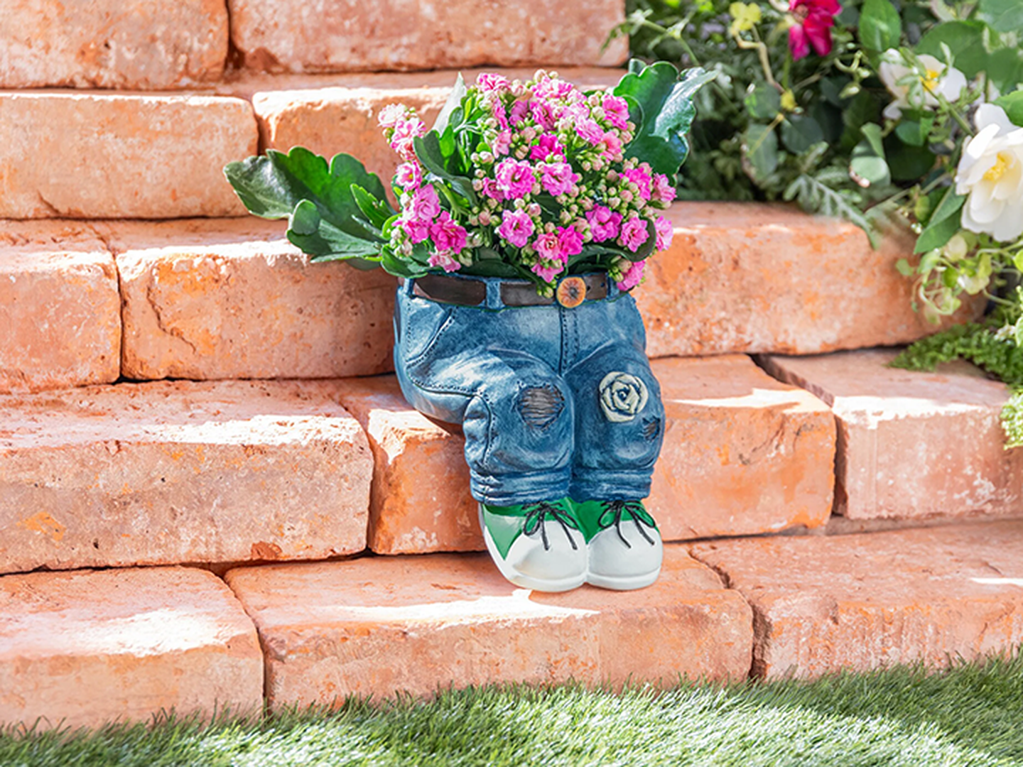 Ein Pflanzgefäß in Form einer Jeanshose, gefüllt mit rosa Blüten und grünen Blättern, steht auf Terrakotta-Stufen inmitten von Grün.