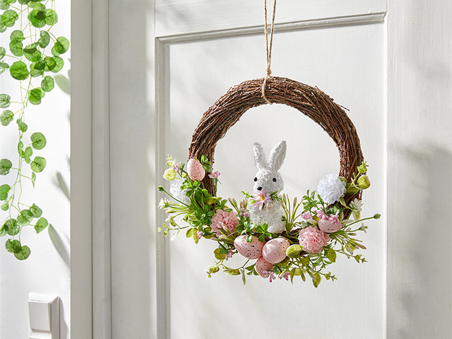 An einer weißen Tür hängt ein Kranz mit einem weißen Kaninchen, rosa und weißen Blumen, grünen Blättern und verzierten Eiern.
