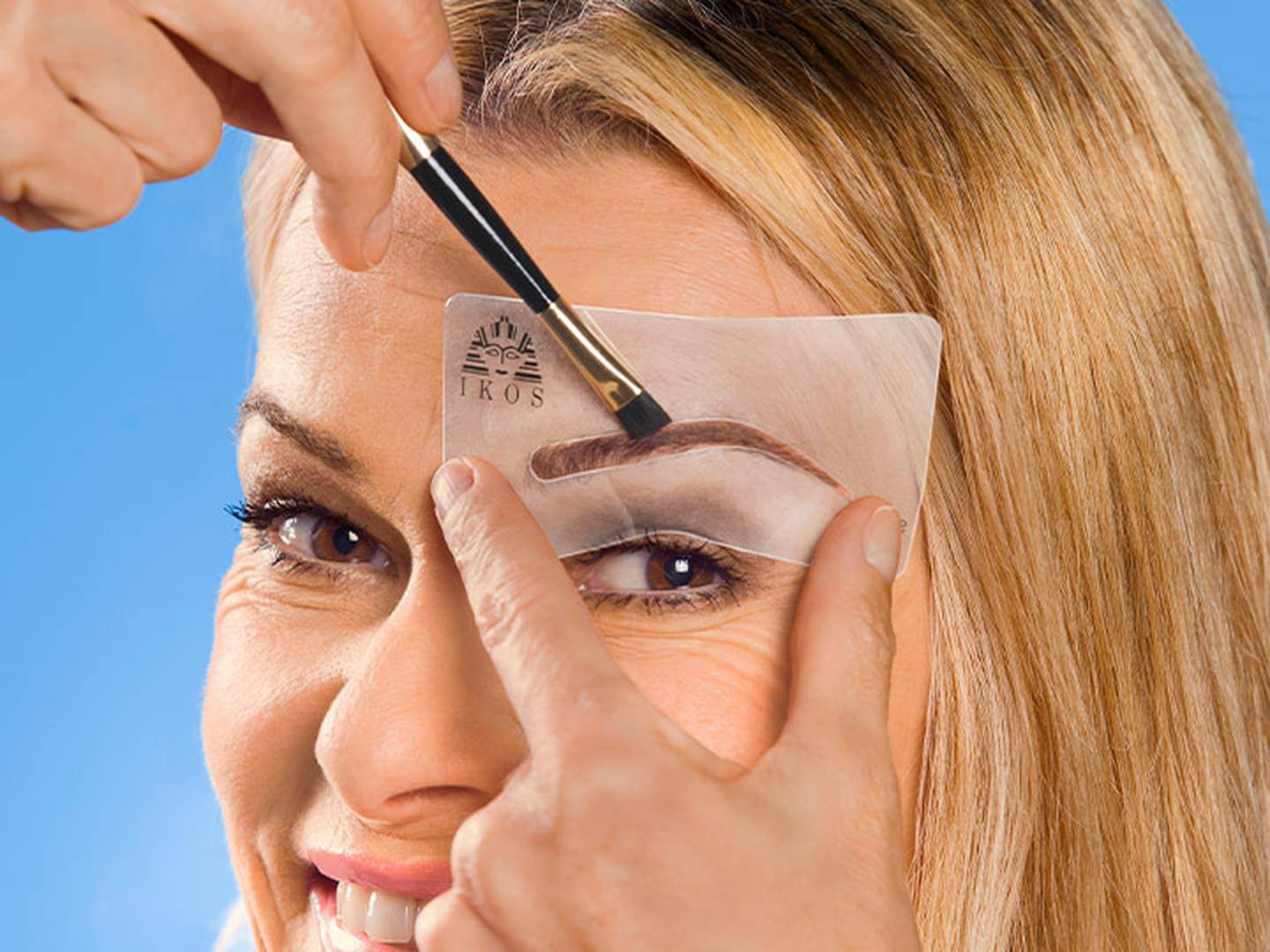 Eine Frau formt mithilfe einer Schablone und eines Pinsels ihre Augenbraue vor einem blauen Hintergrund.