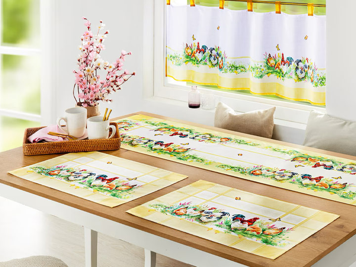 Ein Tisch, gedeckt mit farbenfroher Tischwäsche mit Hahnenmotiven, Kirschblütendekoration und Tassen auf einem Holztablett, steht vor einem Fenster mit passenden Vorhängen.