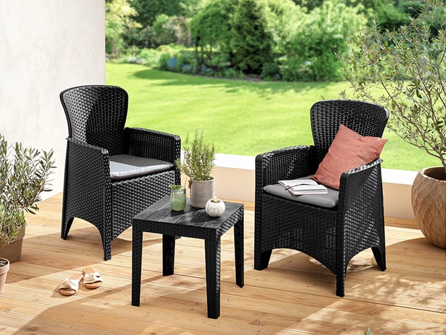 Zwei schwarze Korbstühle mit Kissen auf einer Holzterrasse, ein kleiner Tisch mit Kerzen und Pflanze, mit Blick auf einen üppig grünen Garten.
