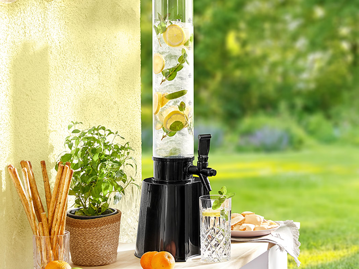 Ein hoher Getränkespender mit Wasser, das mit Zitrone, Limette und Minze aromatisiert ist, steht auf einem Terrassentisch – umgeben von Grissini, einer Topfpflanze und Zitrusfrüchten.