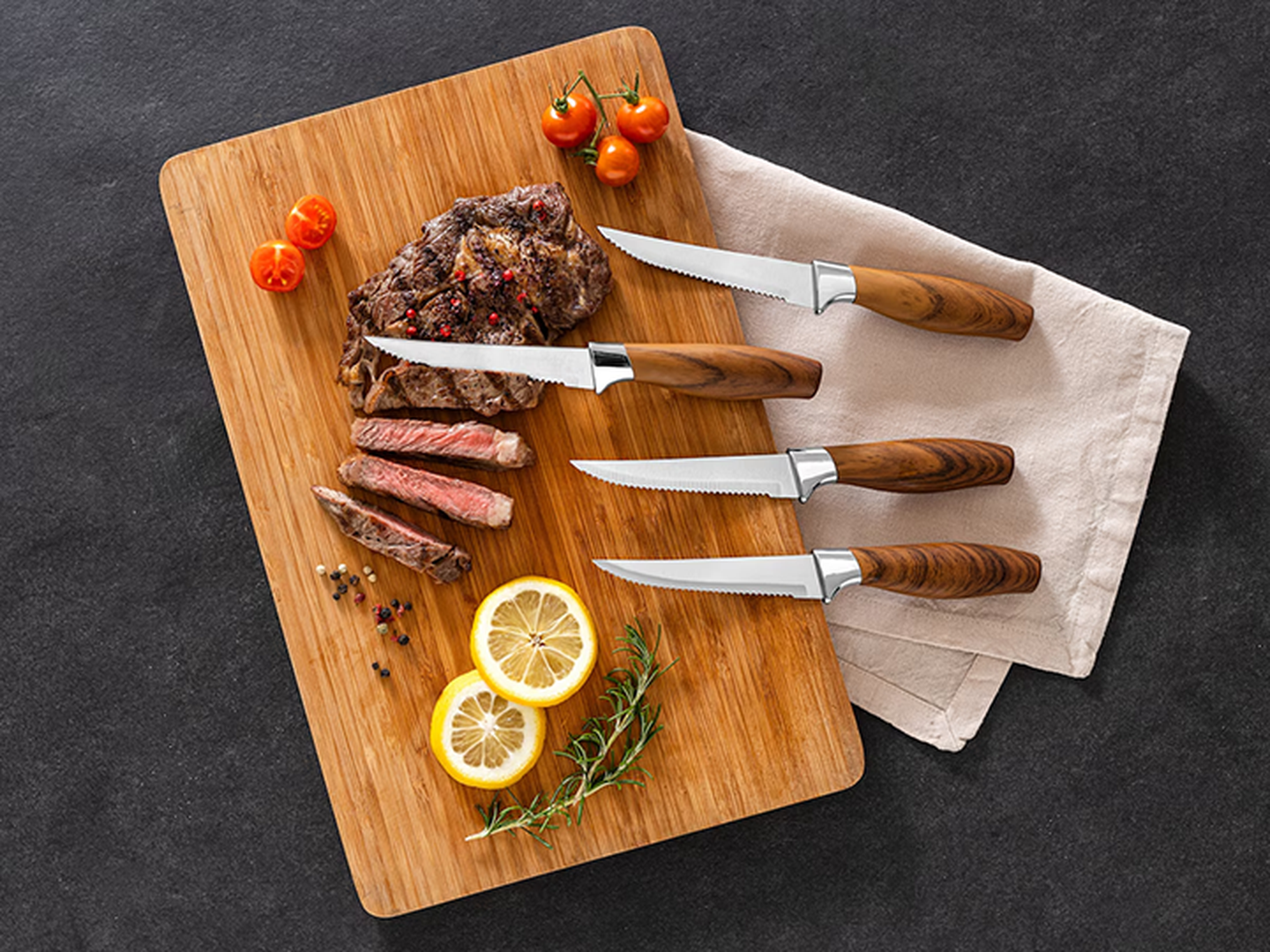 Ein Set Steakmesser auf einem Holzbrett mit geschnittenem Steak, Kirschtomaten, Zitronenscheiben und Kräutern auf einer dunklen Oberfläche.