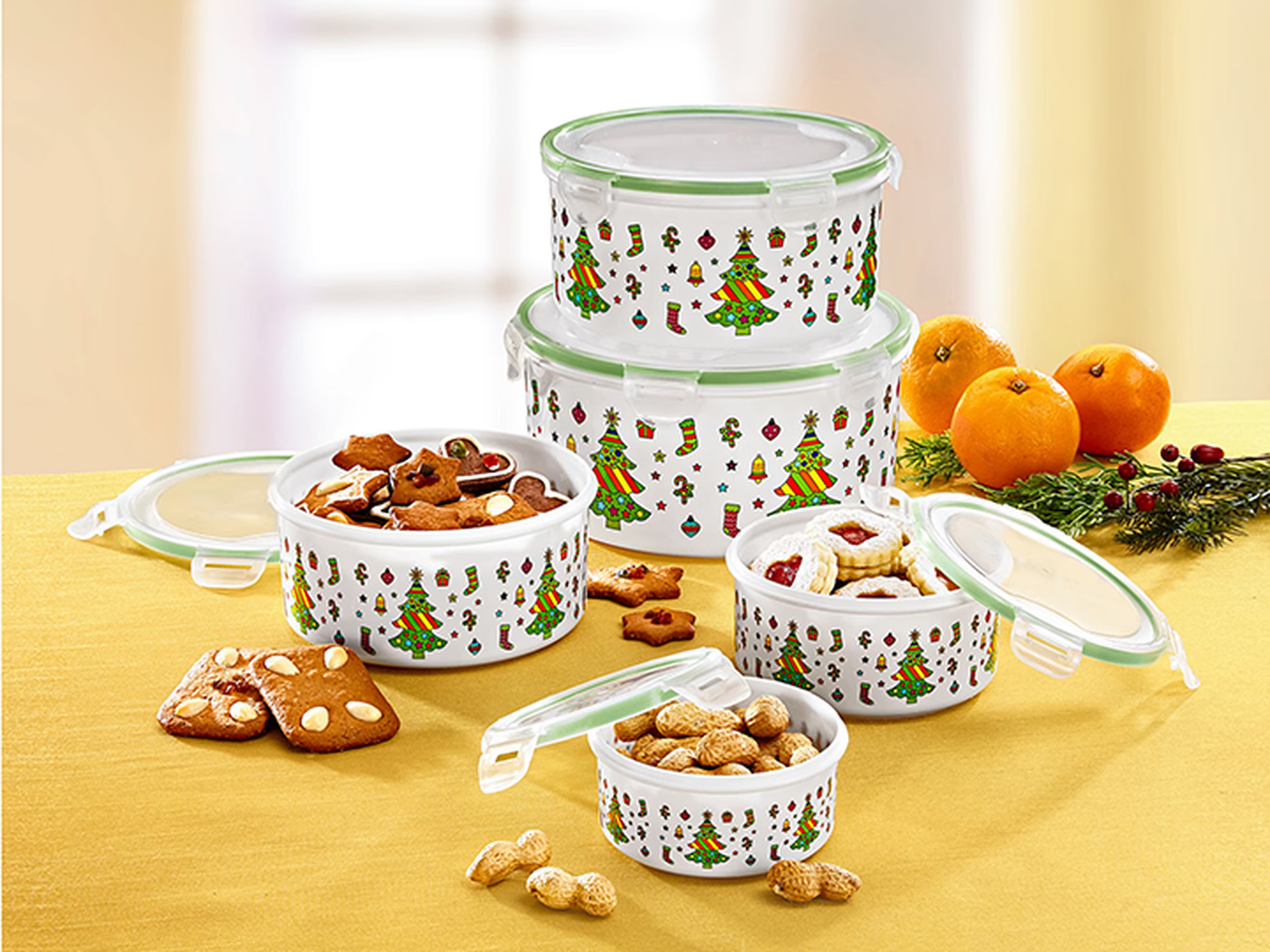 Dekorative Behälter mit Weihnachtsmotiven, gefüllt mit einer Auswahl an Keksen und Erdnüssen, dazu Lebkuchen und Orangen auf einem Tisch.