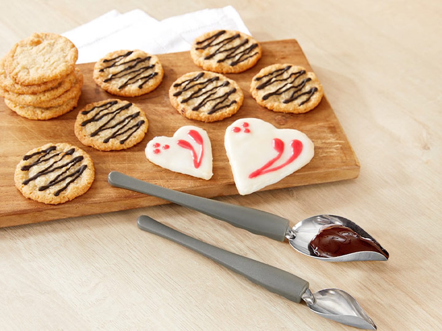 Ein Holzbrett mit runden, mit Schokolade überzogenen Keksen, zwei weiß glasierten Herzkeksen und zwei Löffeln mit geschmolzener Schokolade.