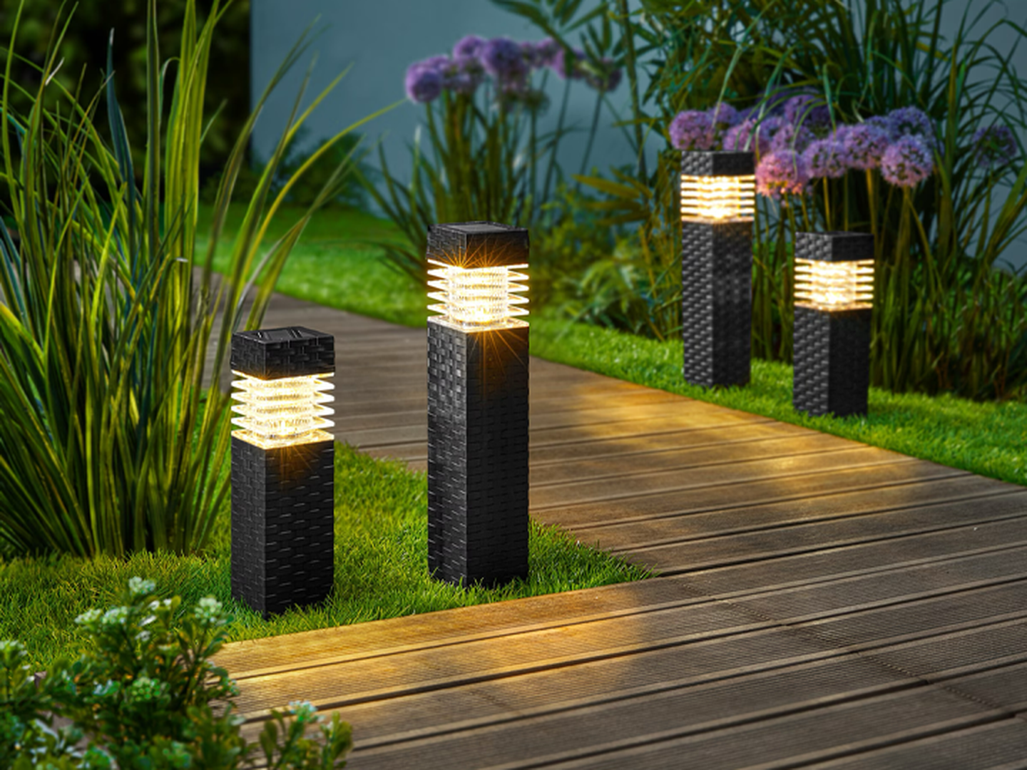 Vier moderne Gartenleuchten beleuchten abends einen Holzsteg, der von üppigem Gras und Pflanzen umgeben ist.