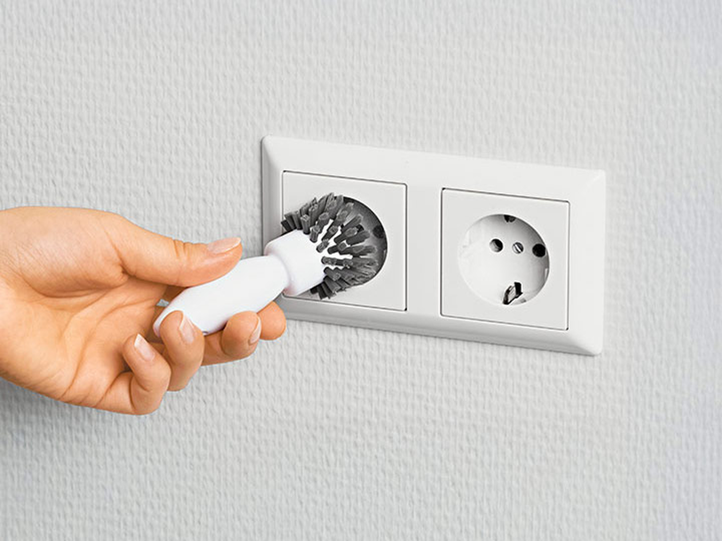 Eine Hand, die eine kleine Bürste hält, reinigt eine Wandsteckdose mit zwei Ausgängen an einer strukturierten weißen Wand.