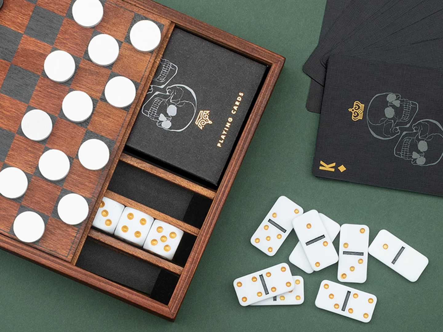 Spielset mit einem hölzernen Schachbrett, weißen Spielsteinen, Würfeln, Spielkarten und Dominosteinen auf grünem Hintergrund.