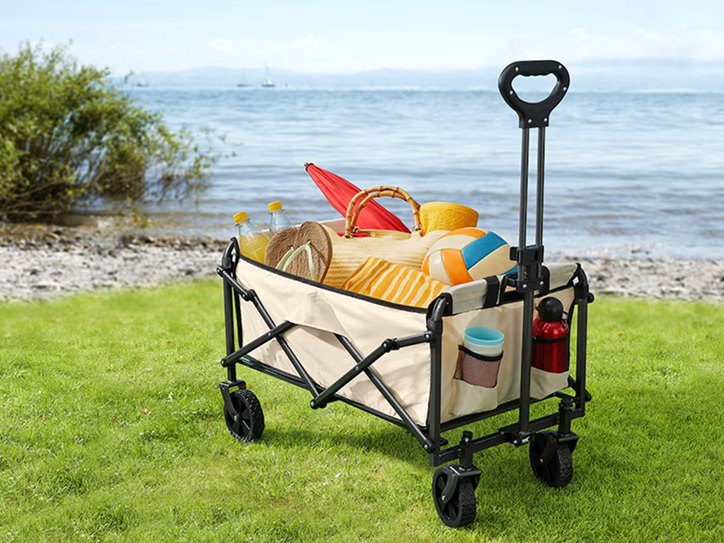 Ein zusammenklappbarer Bollerwagen, gefüllt mit Strandutensilien – darunter Handtücher, ein Hut, ein Ball und Getränke, steht auf der Wiese nahe einem Kiesstrand und ruhigem Wasser.
