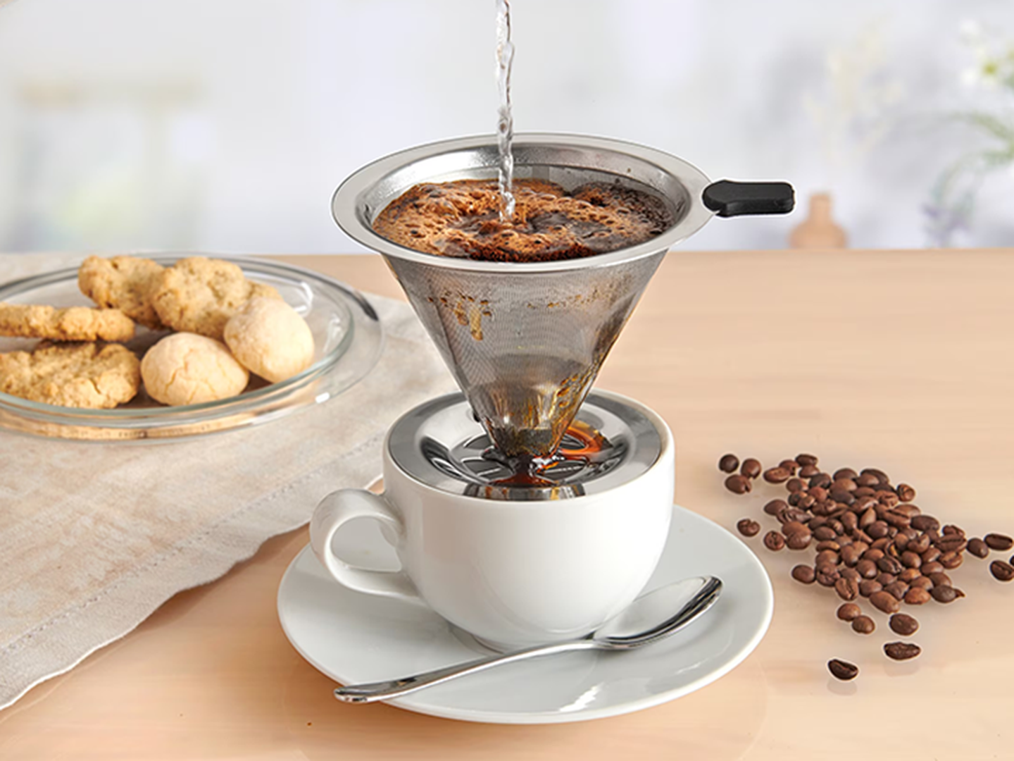 Zubereitung von Pour-Over-Kaffee in einer weißen Tasse mit Metallfilter. Kaffeebohnen und Kekse liegen daneben auf einem Holztisch.