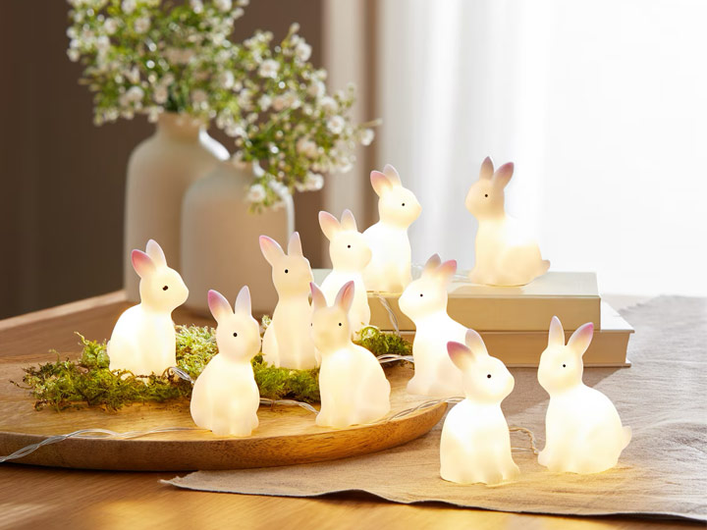 Niedliche, leuchtende Hasenfiguren sind auf einem Tisch mit Grünpflanzen und Büchern arrangiert, in einem sanft beleuchteten Raum mit einer Blumenvase im Hintergrund.