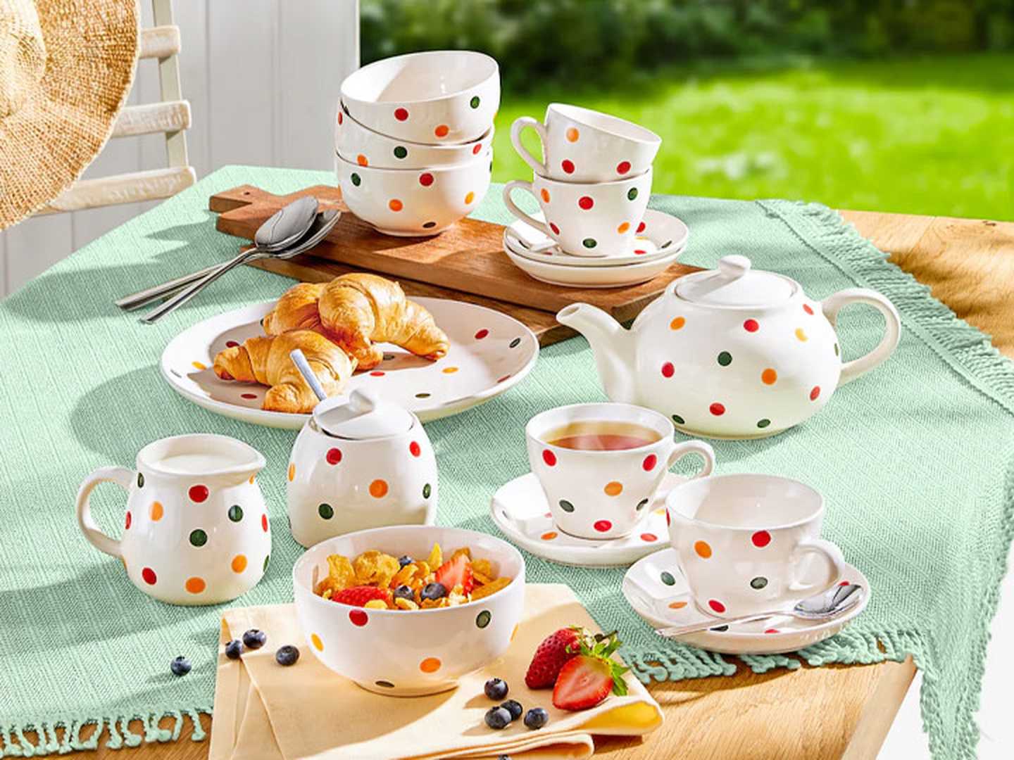 Gepunktetes Teeservice mit Tassen, Untertassen, Teekanne und Tellern auf einer grünen Tischdecke, dazu Croissants, Obst und Müsli.