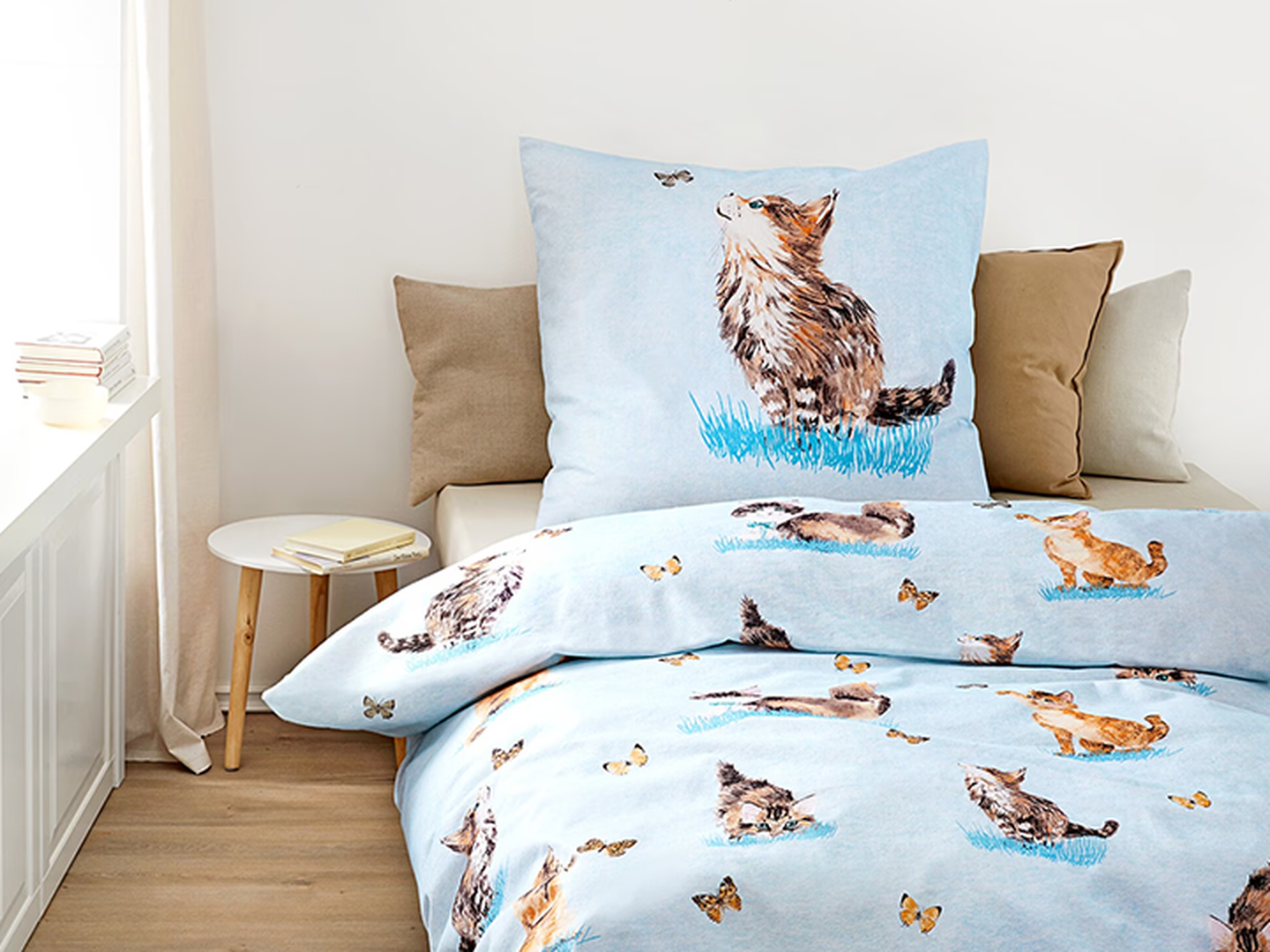 Hellblaues Bettwäsche-Set mit Katzen- und Schmetterlingsmotiven auf einem ordentlich gemachten Bett, ergänzt durch einen kleinen Beistelltisch mit Büchern.