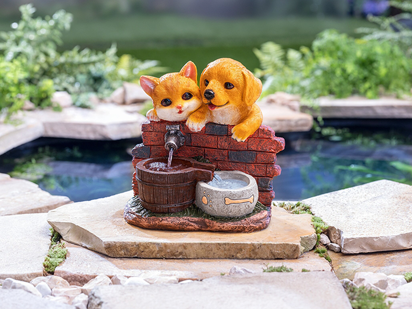 Eine dekorative Figur, die eine Katze und einen Hund zeigt, welche sich an eine Ziegelmauer neben einem kleinen Brunnen auf einer Steinplattform lehnen; im Hintergrund ist Grün zu sehen.