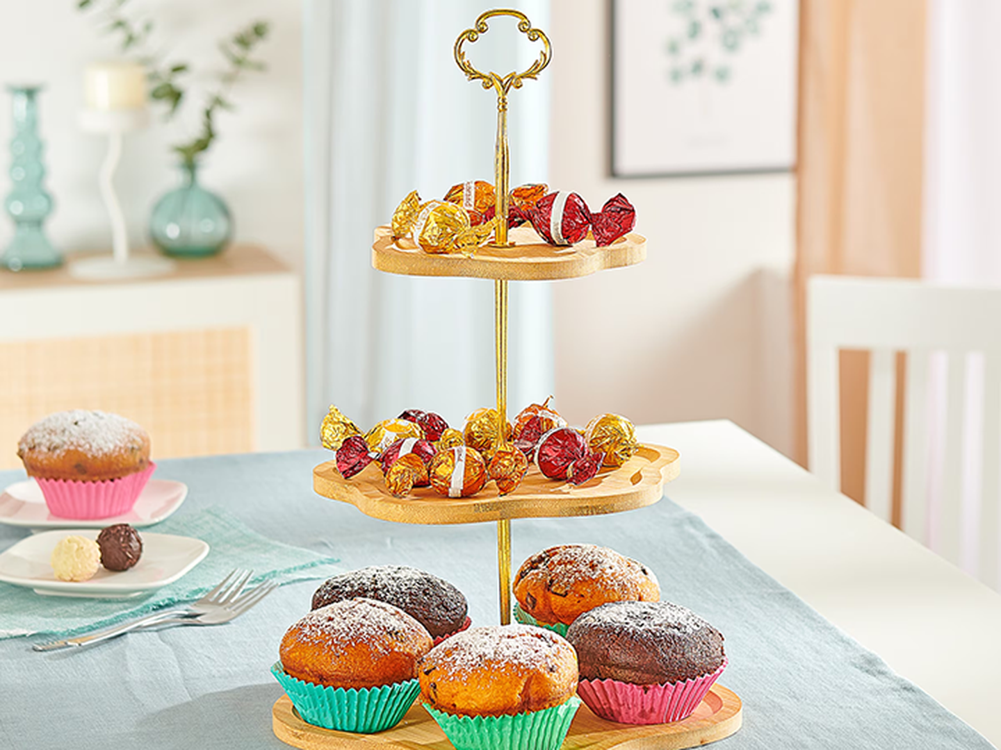 Ein Etageren-Ständer mit eingewickelten Süßigkeiten und mit Puderzucker bestäubten Muffins in bunten Förmchen steht auf einem Tisch, auf dem sich weitere Backwaren und Dekor befinden, in einem hellen Raum.