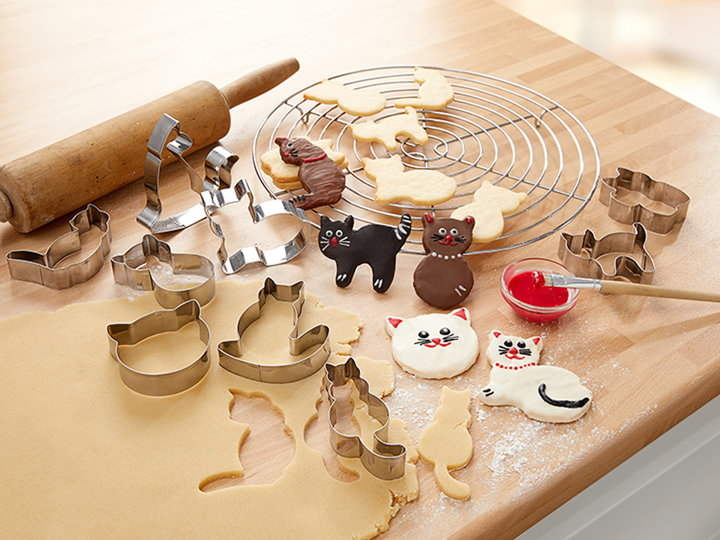 Katzenförmige Kekse mit buntem Zuckerguss auf einem Holztisch. Ausstechformen aus Metall, ein Nudelholz und ein Schälchen mit rotem Zuckerguss stehen daneben.