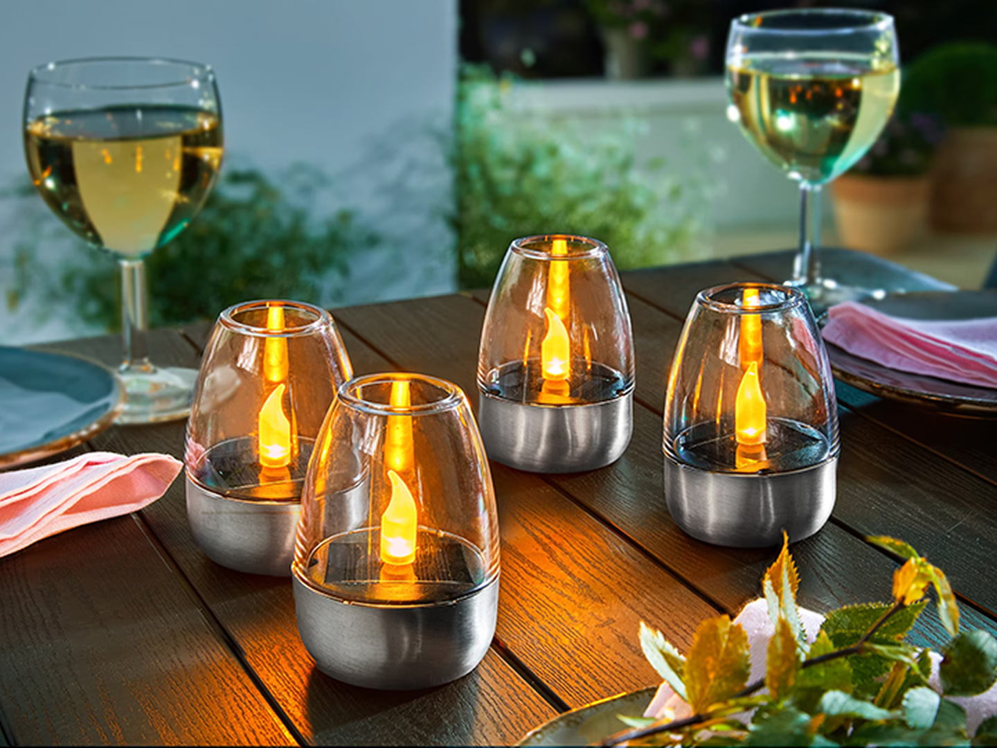 Vier kleine Windlichter auf einem Holztisch, umgeben von Weißweingläsern und rosa Servietten.