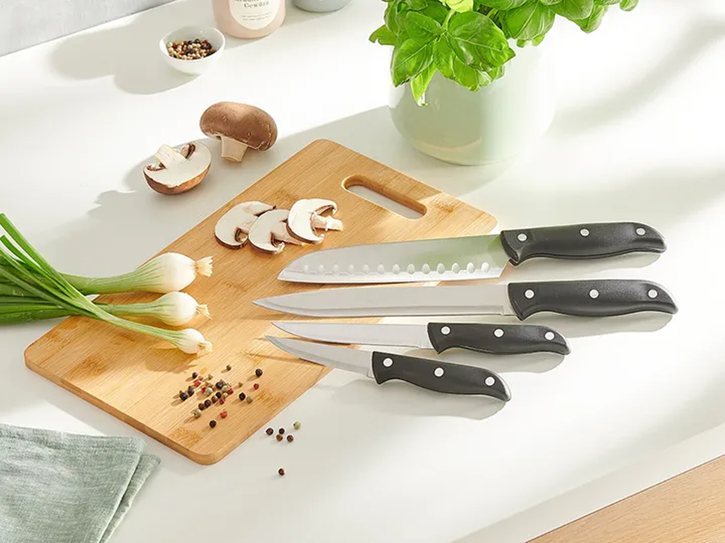 Auf einem Holzschneidebrett liegen vier Küchenmesser mit schwarzen Griffen, daneben geschnittene Champignons, Frühlingszwiebeln und Pfefferkörner, neben einer Basilikumpflanze.
