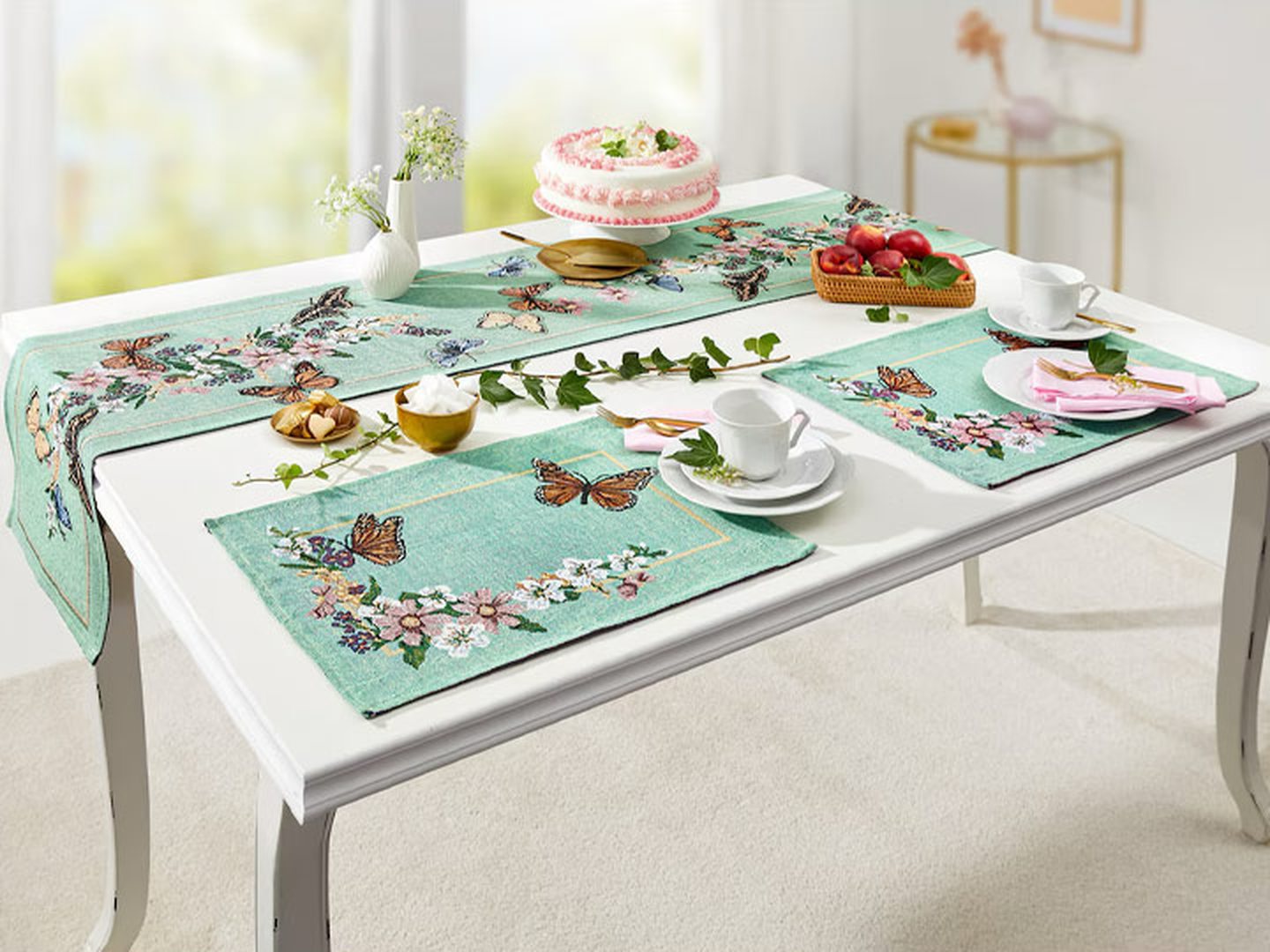 Ein elegant gedeckter Tisch mit Platzsets im Schmetterlingsdesign, Teetassen, einem rosa Kuchen, Kirschen und einer Topfpflanze schafft eine ruhige und beschauliche Atmosphäre beim Essen.