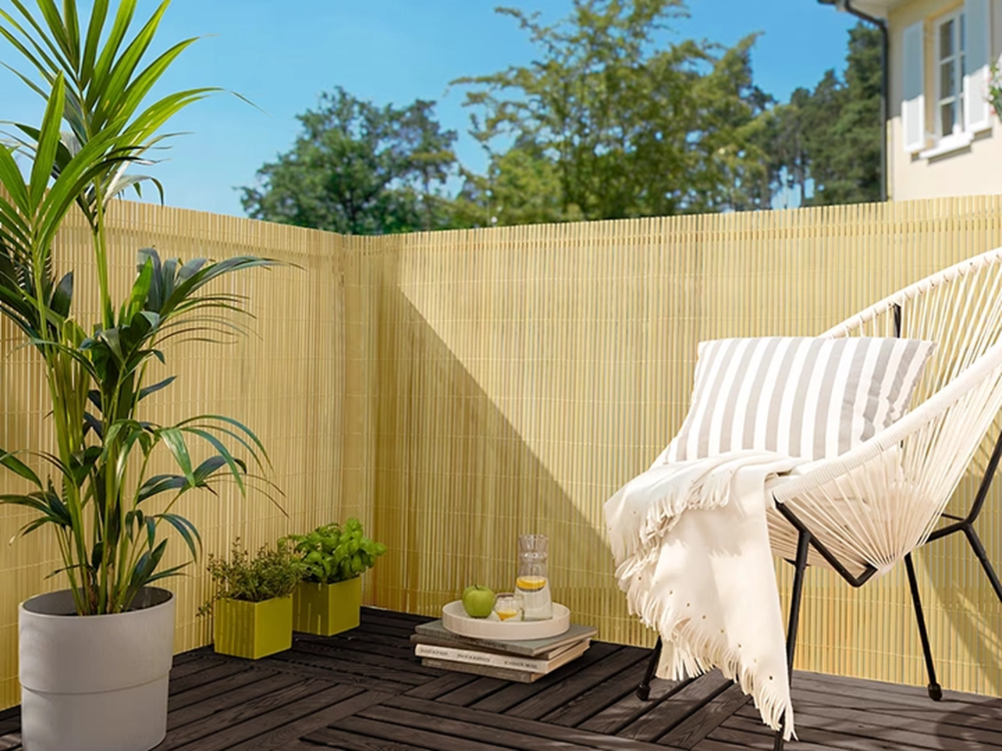 Gemütlicher Balkon mit Korbsessel, gestreiftem Kissen, Topfpflanzen, Holzboden und Bambuszaun unter strahlend blauem Himmel.