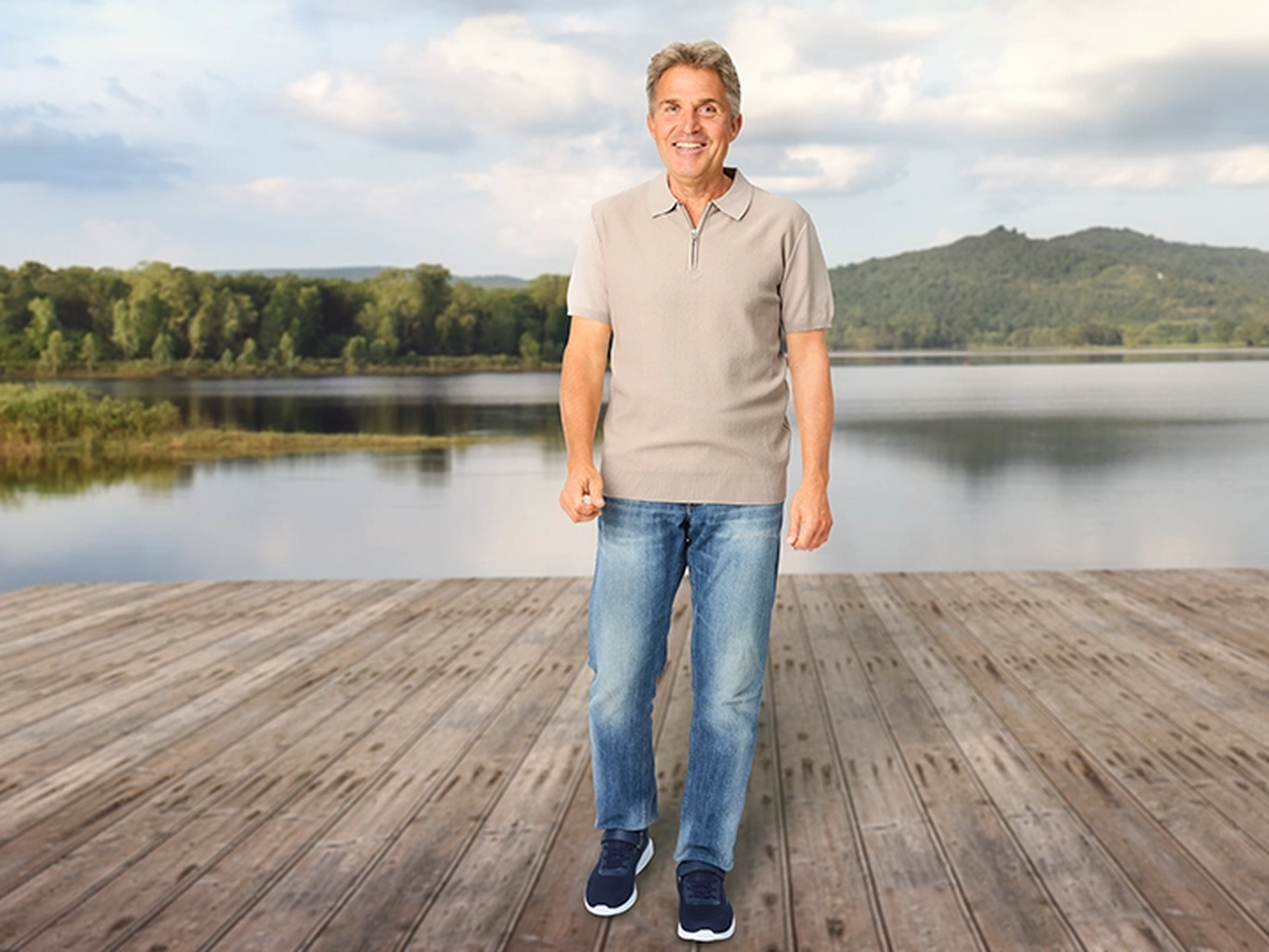 Ein Mann in Jeans und einem legeren Hemd steht lächelnd auf einer Holzterrasse an einem ruhigen See, im Hintergrund sind bewaldete Hügel unter einem bewölkten Himmel zu sehen.