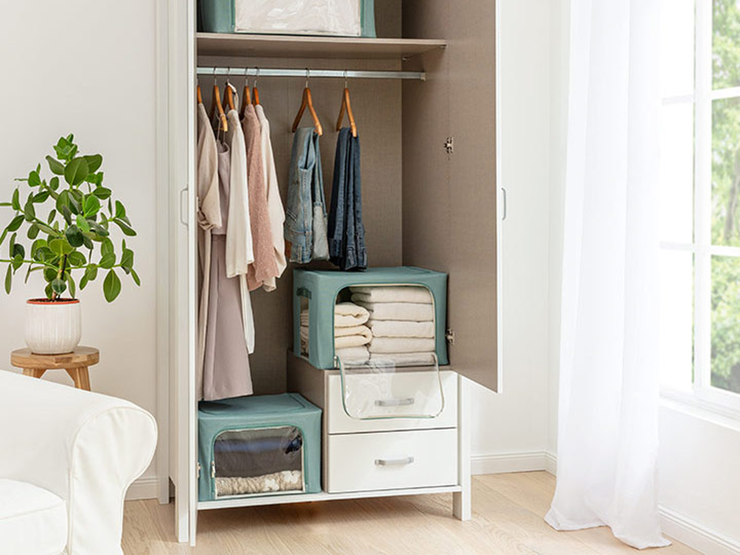 Ein offener Kleiderschrank mit ordentlich sortierter Kleidung, Aufbewahrungsboxen und gefalteter Bettwäsche. Daneben, neben einem hellen Fenster, steht eine Topfpflanze.