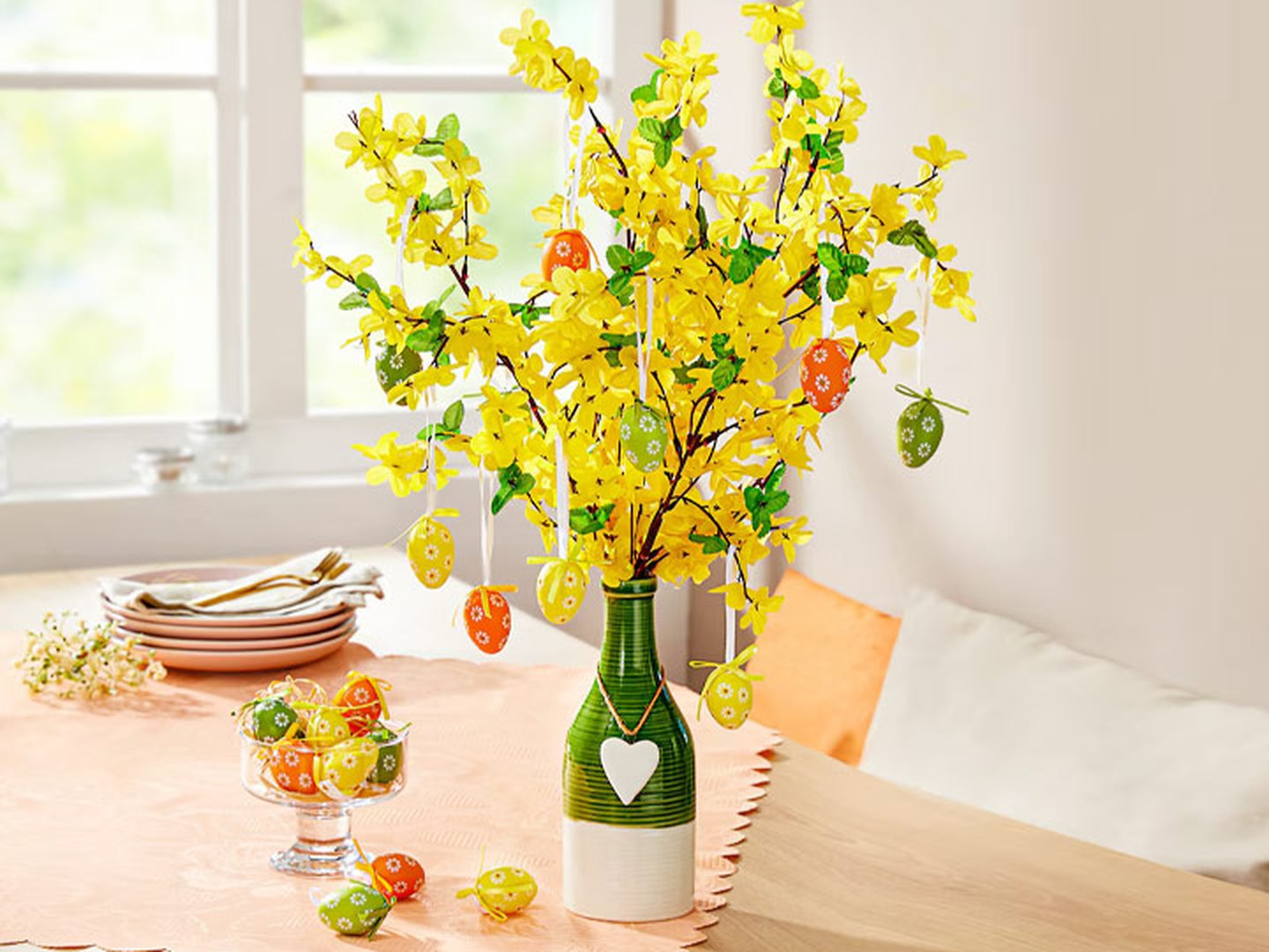 Gelbe blühende Zweige in einer Vase, verziert mit bunten Eierornamenten, auf einem Tisch mit einer Schale mit ähnlichen Eiern und gestapelten Tellern.