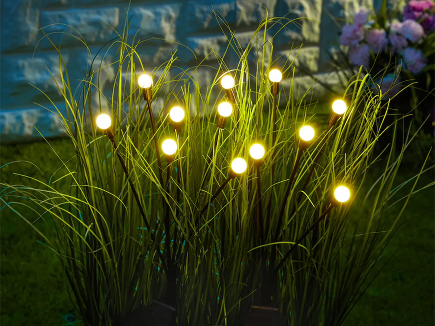 Leuchtende Kugeln auf hohen, grasartigen Stängeln in einem Garten bei Nacht, im Hintergrund eine Steinmauer und Blumen.
